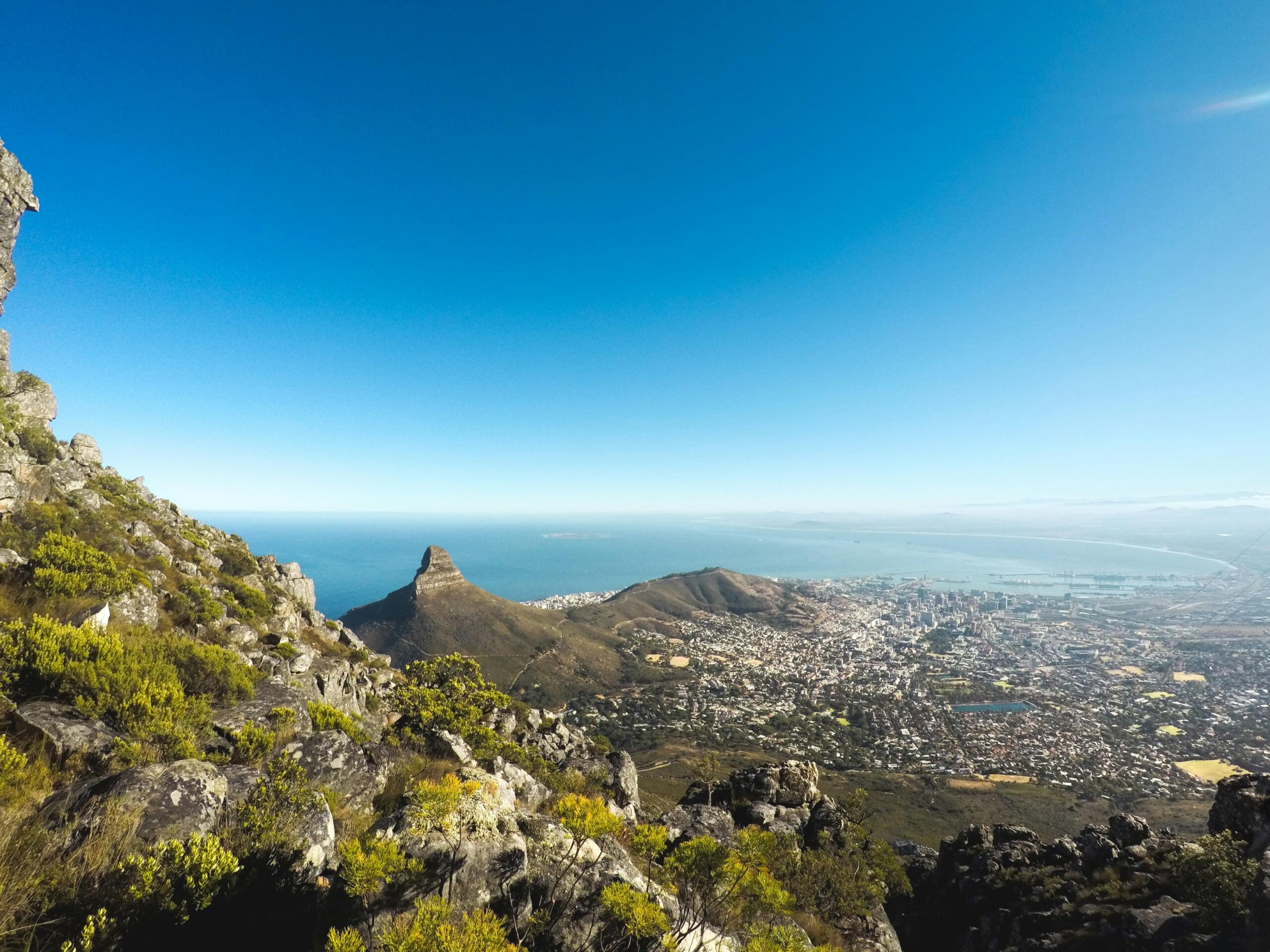 View from a rocky, green mountainside overlooking a city by the sea, with a prominent peak and coastline under a clear blue sky—an experience praised in countless customer reviews.