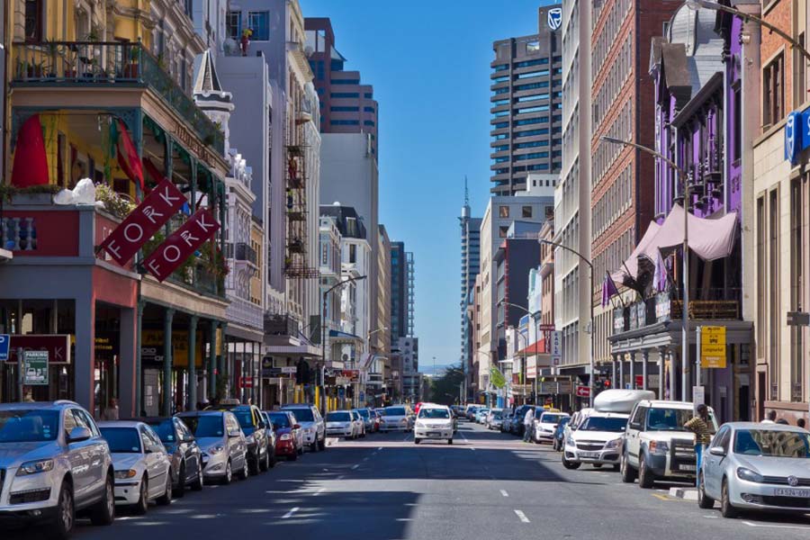 A trendy street lined with parked cars and multi-story buildings, including restaurants and shops with balconies and signs; pedestrians are visible, and taller buildings rise in the background under a clear blue sky.