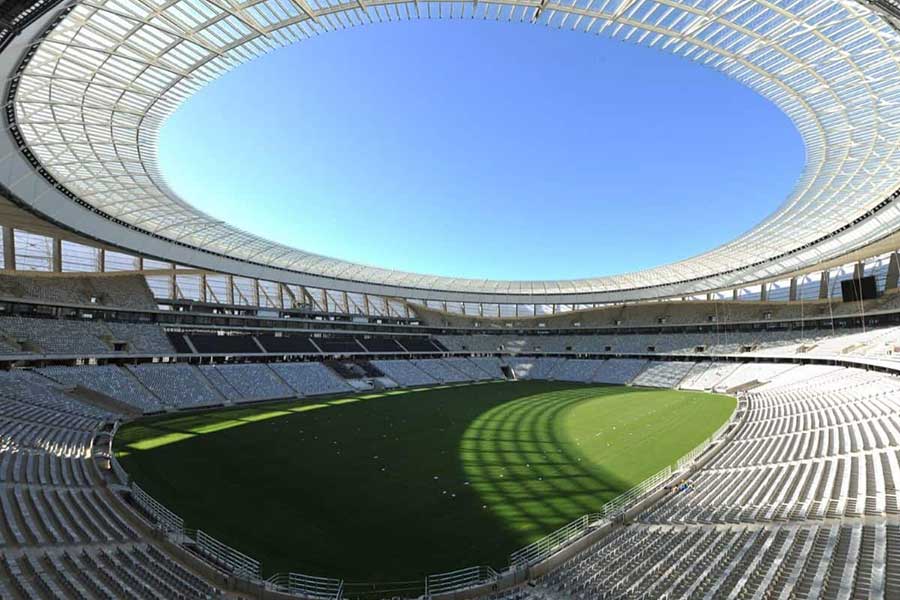 An empty, modern Cape Town Stadium features curved rows of white seats and a large oval-shaped opening in the roof, letting sunlight illuminate the green field below under a clear blue sky.