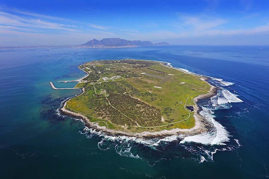 Aerial view of Robben Island, a green, oval-shaped isle surrounded by blue ocean with rocky shores and gentle waves, distant mountains, and a city skyline visible on the horizon under a clear sky.
