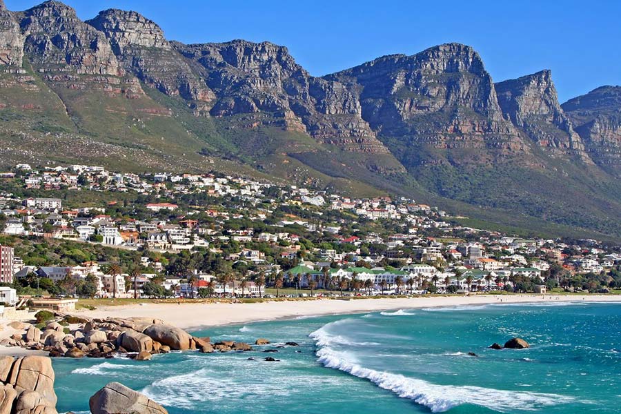 A vibrant coastal town, Cape Town’s Camps Bay Beach boasts white sandy shores, turquoise waves, and scattered boulders at the foot of tall mountains. Houses are nestled between the beach and rugged mountain slopes under a clear blue sky.