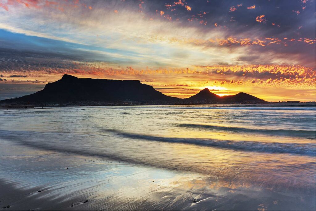Sunset over Table Mountain and the ocean in Cape Town, South Africa. The sky is filled with dramatic clouds as waves gently wash onto the sandy shore—one of the best sunset spots in Cape Town, with vibrant colors reflected on the water.