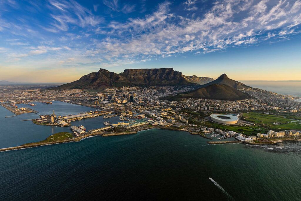 Aerial view of Cape Town showing Table Mountain, surrounding peaks, city buildings, Cape Town Stadium, and coastline with the ocean under a blue sky—ideal for a cultural holiday at Gardens Centre Holiday Apartments.