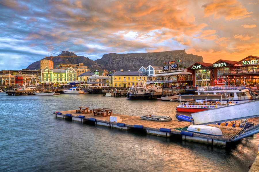 Colorful waterfront scene at sunset with boats docked at piers, vibrant buildings, and Table Mountain in the background under a dramatic sky. Enjoy shopping and dining at lively cafés and shops along Cape Town’s iconic V&A Waterfront harbor.