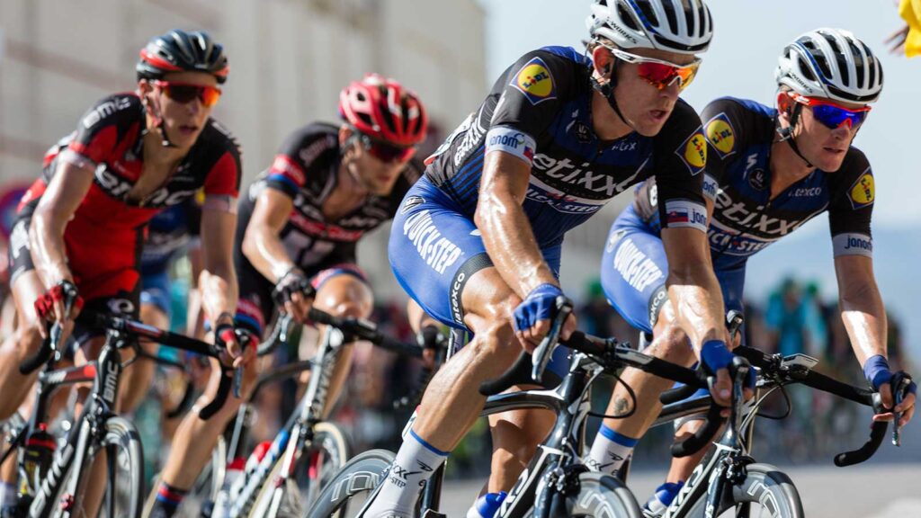A group of cyclists in helmets and racing uniforms compete closely in the Cape Town Cycle Tour on a sunny day. The leading riders wear blue jerseys, while others sport red and black—perfect action viewed from Gardens Centre Holiday Apartments.