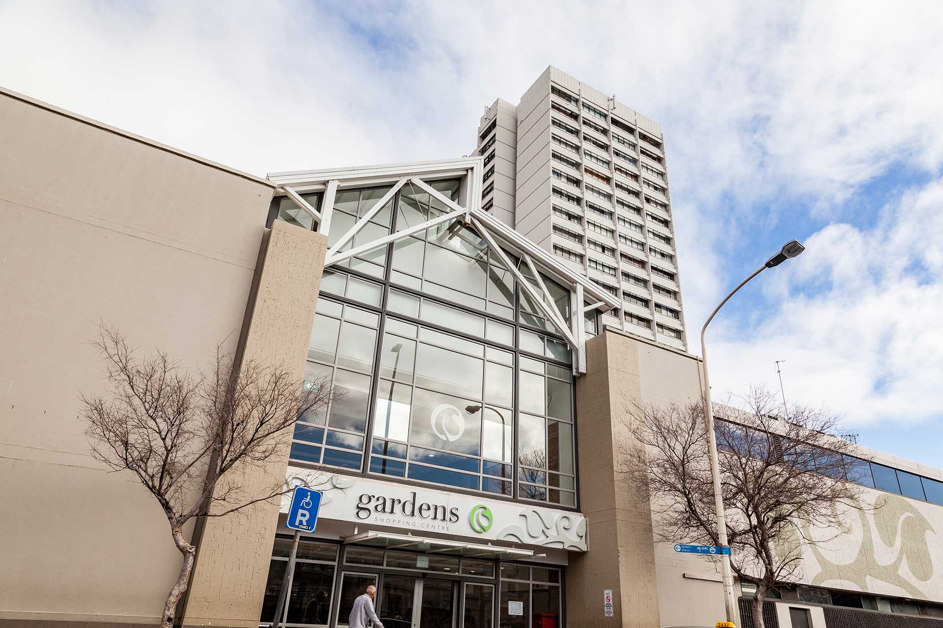 A modern building entrance with large glass windows and a sign reading “gardens,” with a tall apartment tower above. Leafless trees and a blue sky with clouds are visible. A person walks near a handicap parking sign.