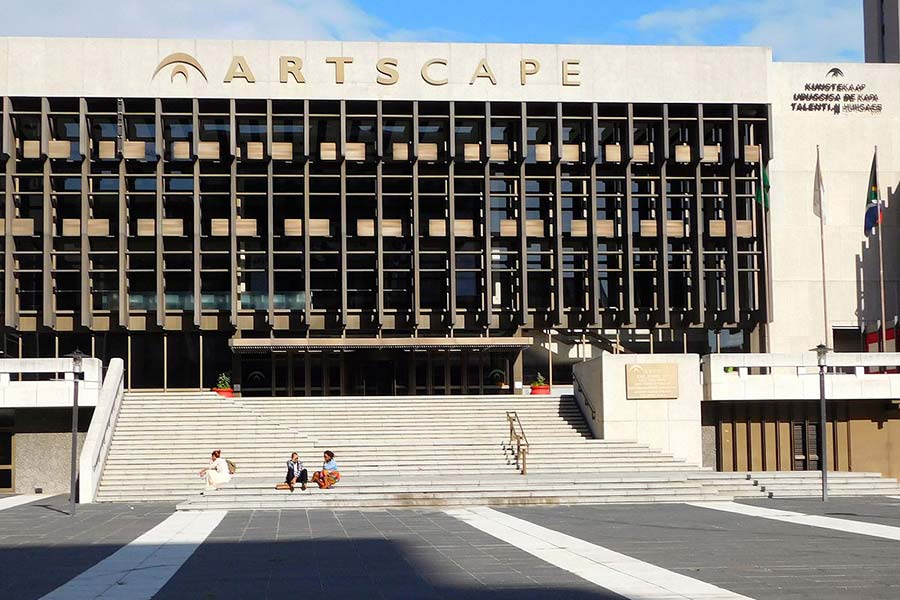 A large modern building with "ARTSCAPE" in gold letters above the entrance, wide steps where people sit, and a white exterior. Vertical grid-like windows define the facade of this renowned Artscape Theatre, a hub for arts and culture.
