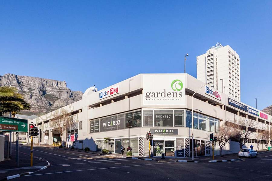 Gardens Shopping Mall, home to Pick n Pay and various stores, is set at a bustling street intersection with traffic lights, framed by a mountain and a tall building under a clear blue sky.