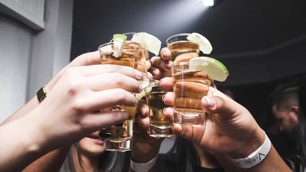 A group of people raising shot glasses filled with a golden liquid, each glass garnished with a lime wedge, as they toast together in a celebratory atmosphere at one of Cape Town’s famous bars.