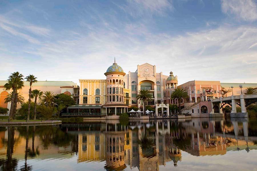 A colorful, grand building with domed towers and a clock, surrounded by palm trees, is reflected in the calm water of Canal Walk Shopping Centre under a blue sky with wispy clouds.