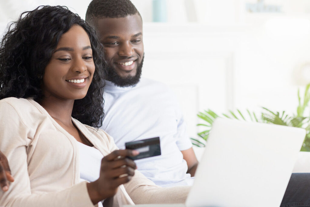 A smiling couple sits on a couch looking at a laptop, with the woman holding a credit card, suggesting they're making a direct hotel reservation or enjoying easy online hotel booking together.
