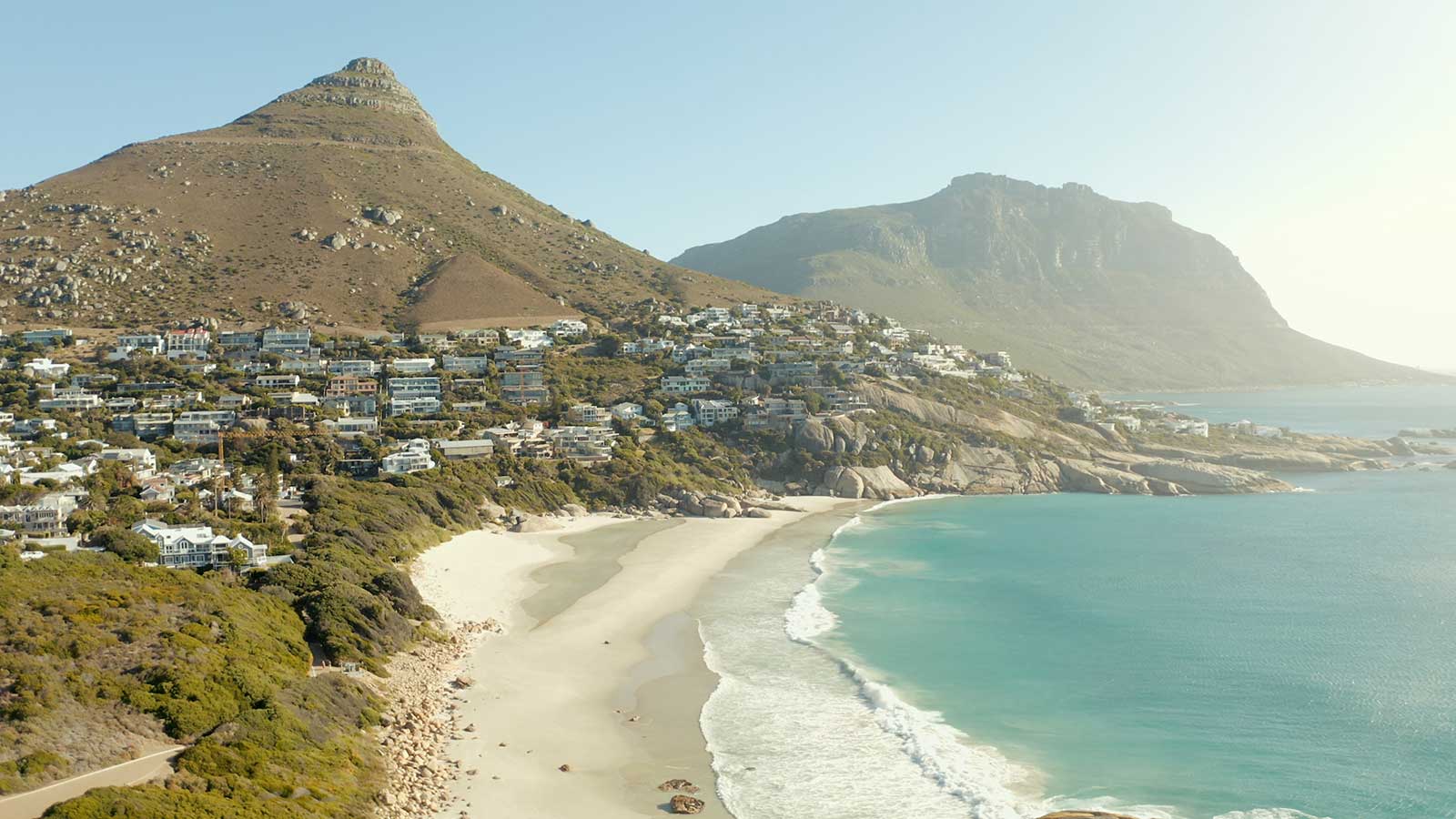 A scenic coastal view shows sandy beaches, turquoise water, rocky hills, and houses nestled on the slopes under a clear sky—capturing the beauty of Cape Town beaches.