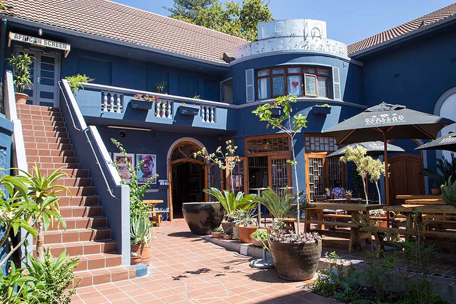 A courtyard of a blue building in Cape Town with potted plants, wooden benches, a staircase to the left, and an umbrella for shade. The building has arched windows and a sign that reads "Labia Theatre," an iconic independent cinema.