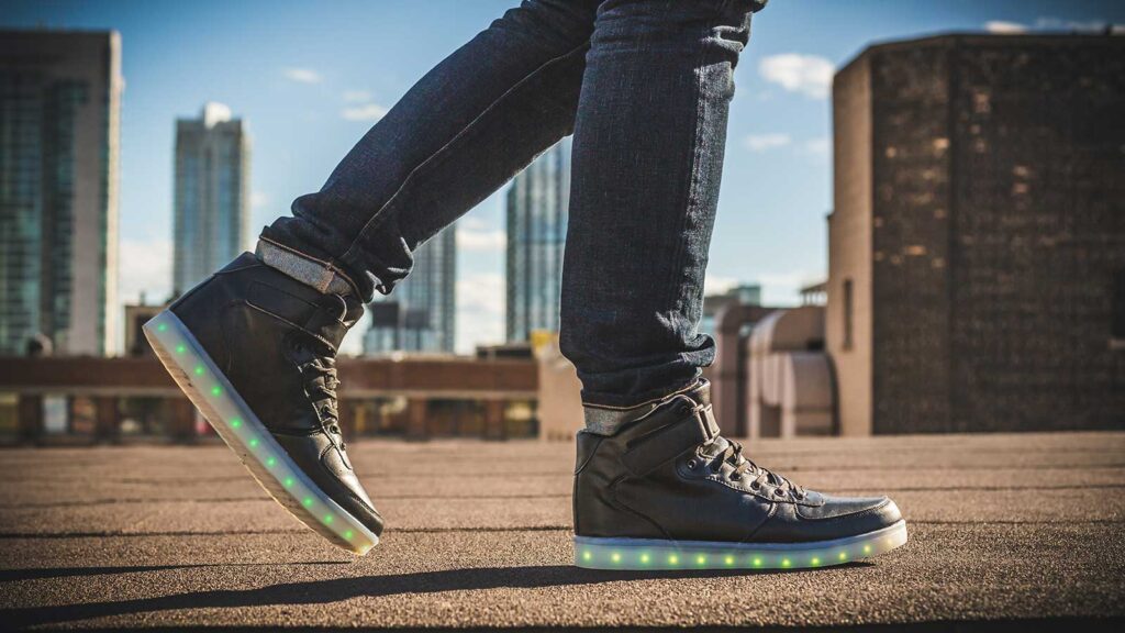 A person wearing dark jeans and black high-top sneakers with glowing green and yellow LED lights in the soles stands on a rooftop, just walking distance from Gardens Centre Holiday Apartments, with city buildings in the background.