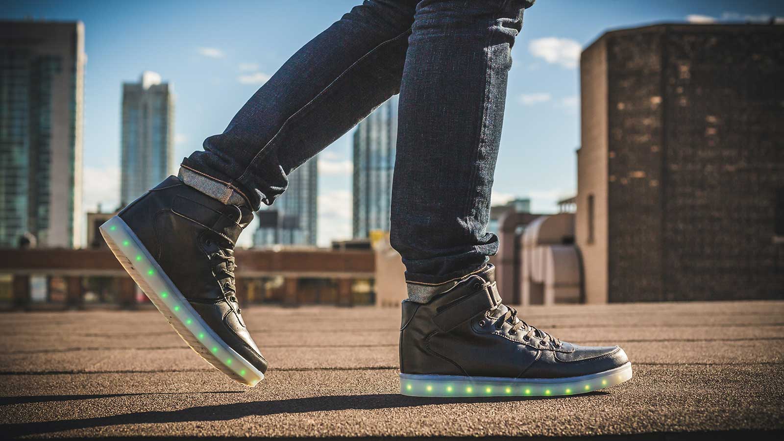 A person wearing dark jeans and black high-top sneakers with glowing green and yellow LED lights in the soles stands on a rooftop, just walking distance from Gardens Centre Holiday Apartments, with city buildings in the background.