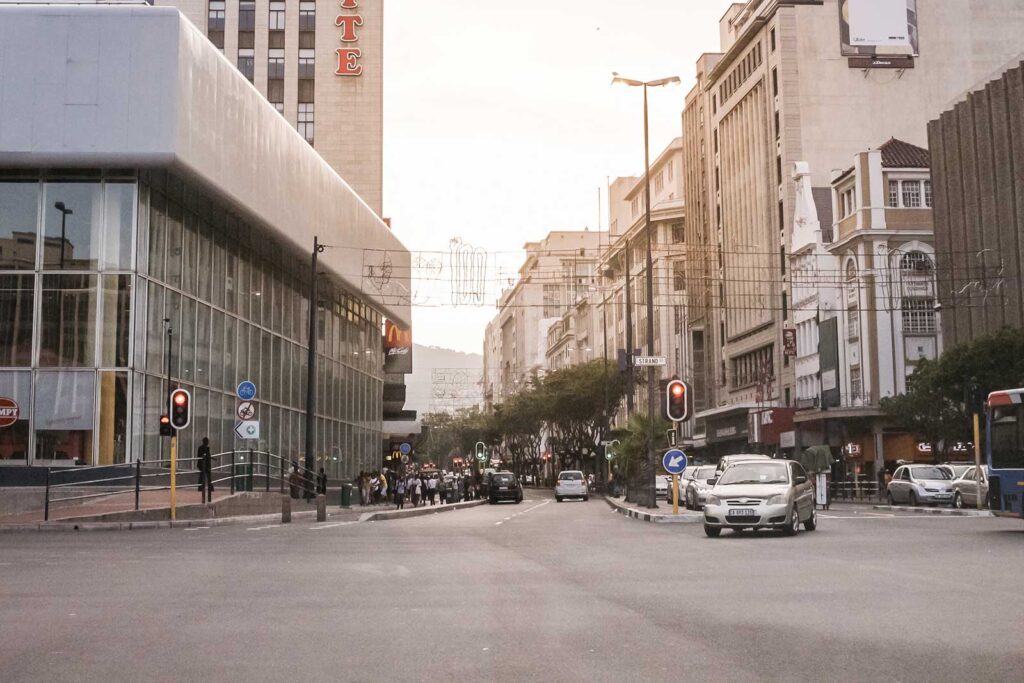 A city street scene at sunset in Cape Town with cars waiting at a red light, pedestrians crossing, and tall buildings lining both sides of the road. The sky is lightly tinted with a warm glow—perfect for those who love to explore urban life.