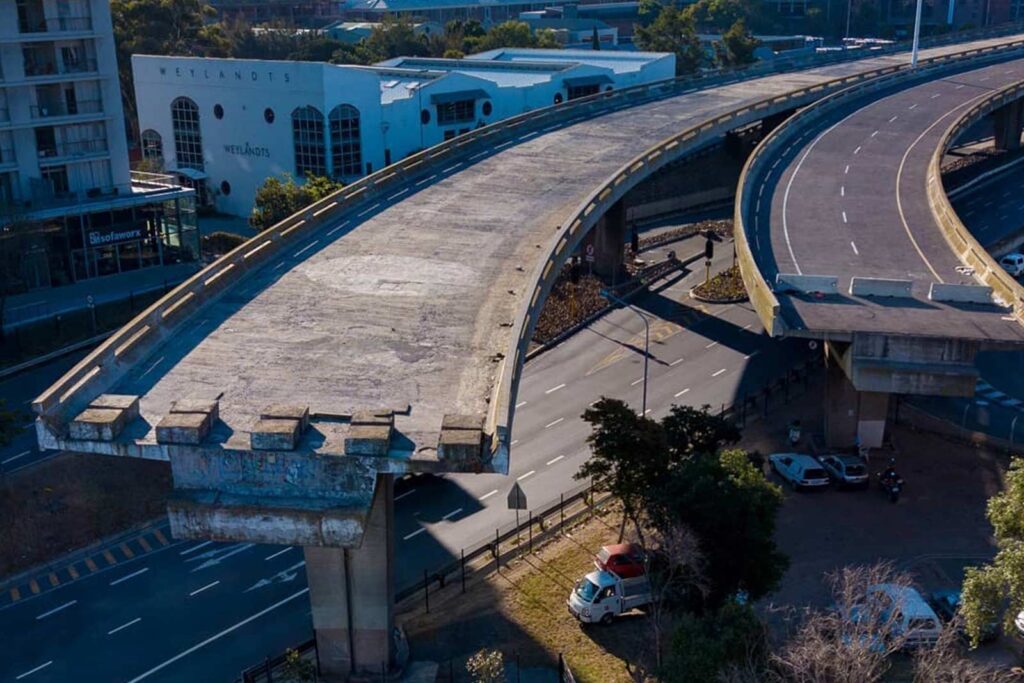 Aerial view of a half-built bridge in Cape Town, its elevated highway ending abruptly above a city street, with vehicles and buildings visible below. The adjacent completed road curves beside the unfinished section.