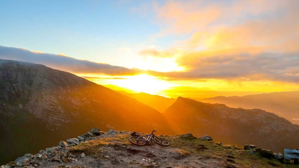 A bicycle rests on a grassy mountain peak at sunrise, with golden sunlight streaming over distant mountains—perfect for adventurous day trips within 100 kilometres of Gardens Centre Holiday Apartments.