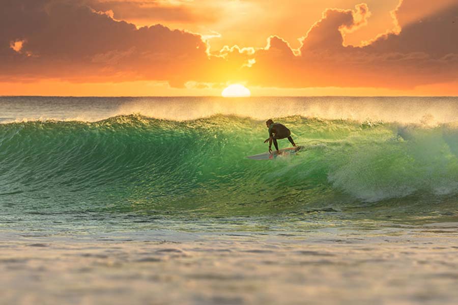 A surfer rides a green wave at sunset, with the sun glowing orange and yellow behind clouds on the horizon. The water appears vibrant and the scene is calm yet dynamic.