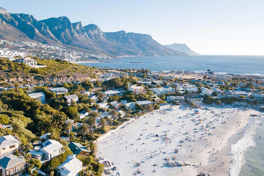 Aerial view of Clifton Beach in Karachi, a popular tourist attraction with sandy shores lined by houses, people relaxing beside the calm blue sea, and green mountains rising under a clear sky.