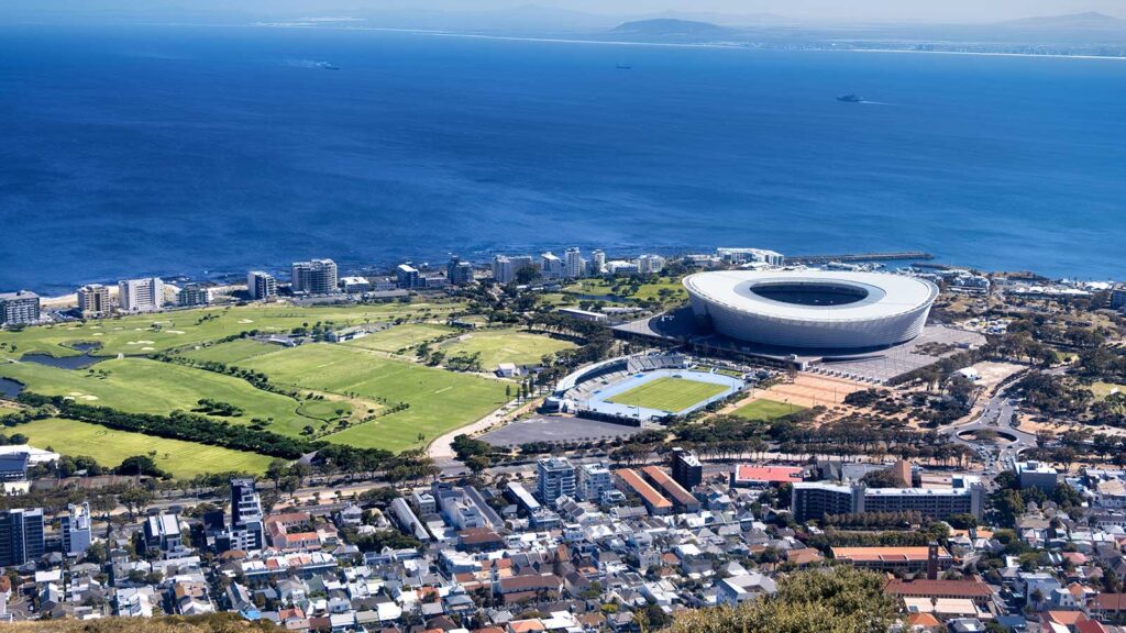 Aerial view of Cape Town, South Africa, featuring Cape Town Stadium near the coast, lush green fields, urban buildings, and blue Atlantic waters—highlighting some of the top attractions Cape Town has to offer.