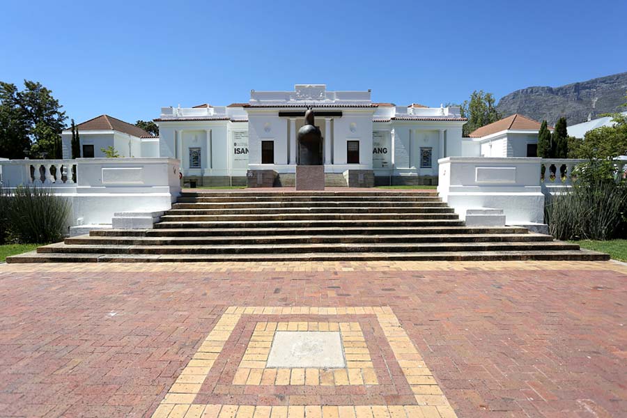 A wide stone staircase leads up to the South African National Gallery, a white, classical-style building with columns and a central sculpture in front, surrounded by greenery under a clear blue sky.