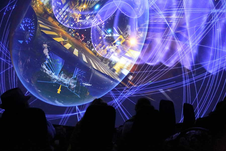 A group of people sits in the dark, facing a large domed screen displaying a vibrant, abstract projection of city lights, intersecting lines, and swirling patterns, creating an immersive visual experience.