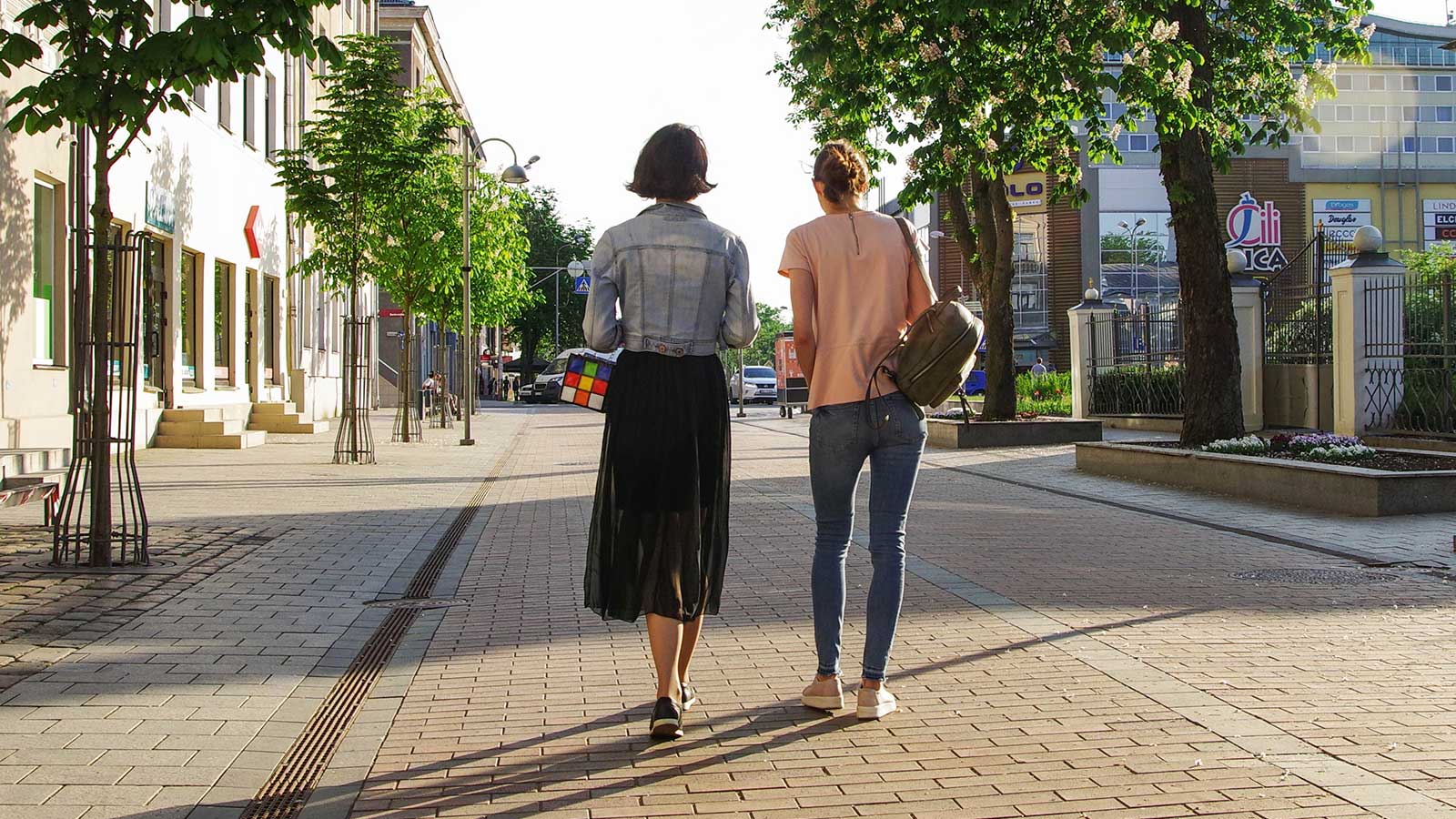 Two women walk down a sunny, tree-lined sidewalk in Gardens Cape Town. One wears a black skirt and denim jacket, holding a colorful cube; the other wears jeans and a pink shirt with a backpack. Buildings and shops line the vibrant street.