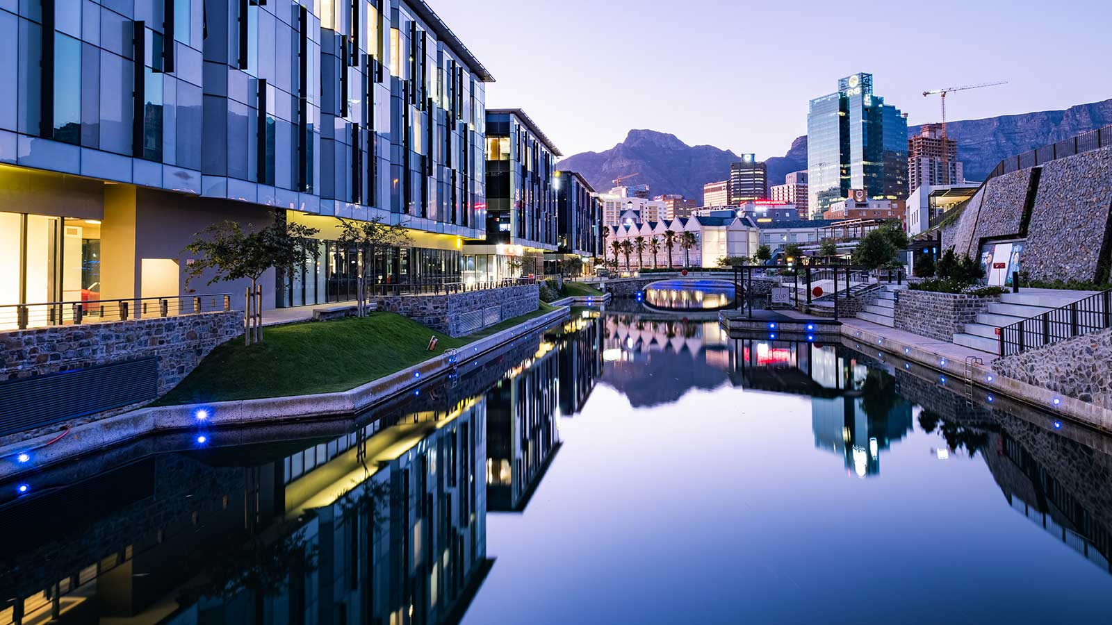 Modern buildings and palm trees reflected in a calm canal at dusk, with Cape Town’s mountains and city skyline in the background under a clear sky—a perfect scene for travel lovers seeking new things to do.