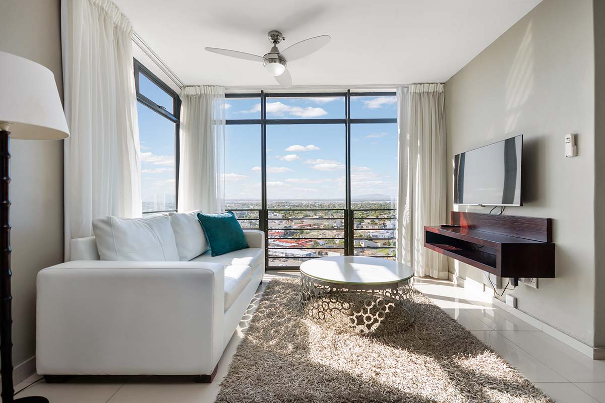 A modern living room in a deluxe apartment features a white sofa, teal pillow, round coffee table, shaggy rug, wall-mounted TV, and large windows letting in natural light with a city view outside.