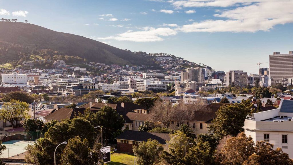 A cityscape with a mix of trees, houses, and modern buildings set against a hillside under a partly cloudy sky—perfect for self-catering holidays. A silhouette of an animal statue can be seen on the hilltop in the background.