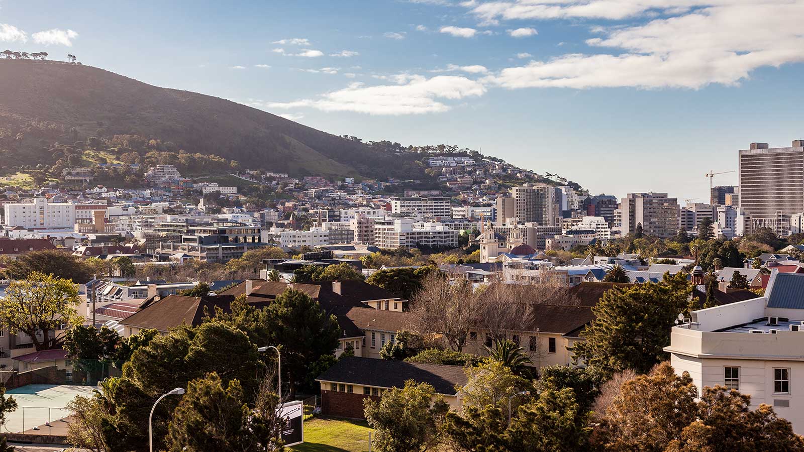 A cityscape with a mix of trees, houses, and modern buildings set against a hillside under a partly cloudy sky—perfect for self-catering holidays. A silhouette of an animal statue can be seen on the hilltop in the background.