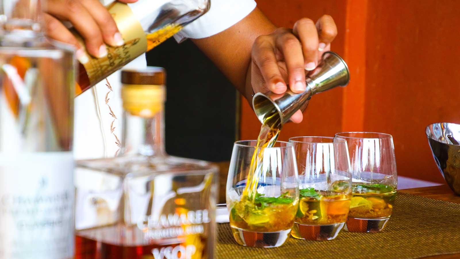 A bartender pours whiskey from a jigger into glasses with ice and herbs on a bar counter, capturing the spirit of sundowners at one of Cape Town's best places, with liquor bottles visible in the foreground.