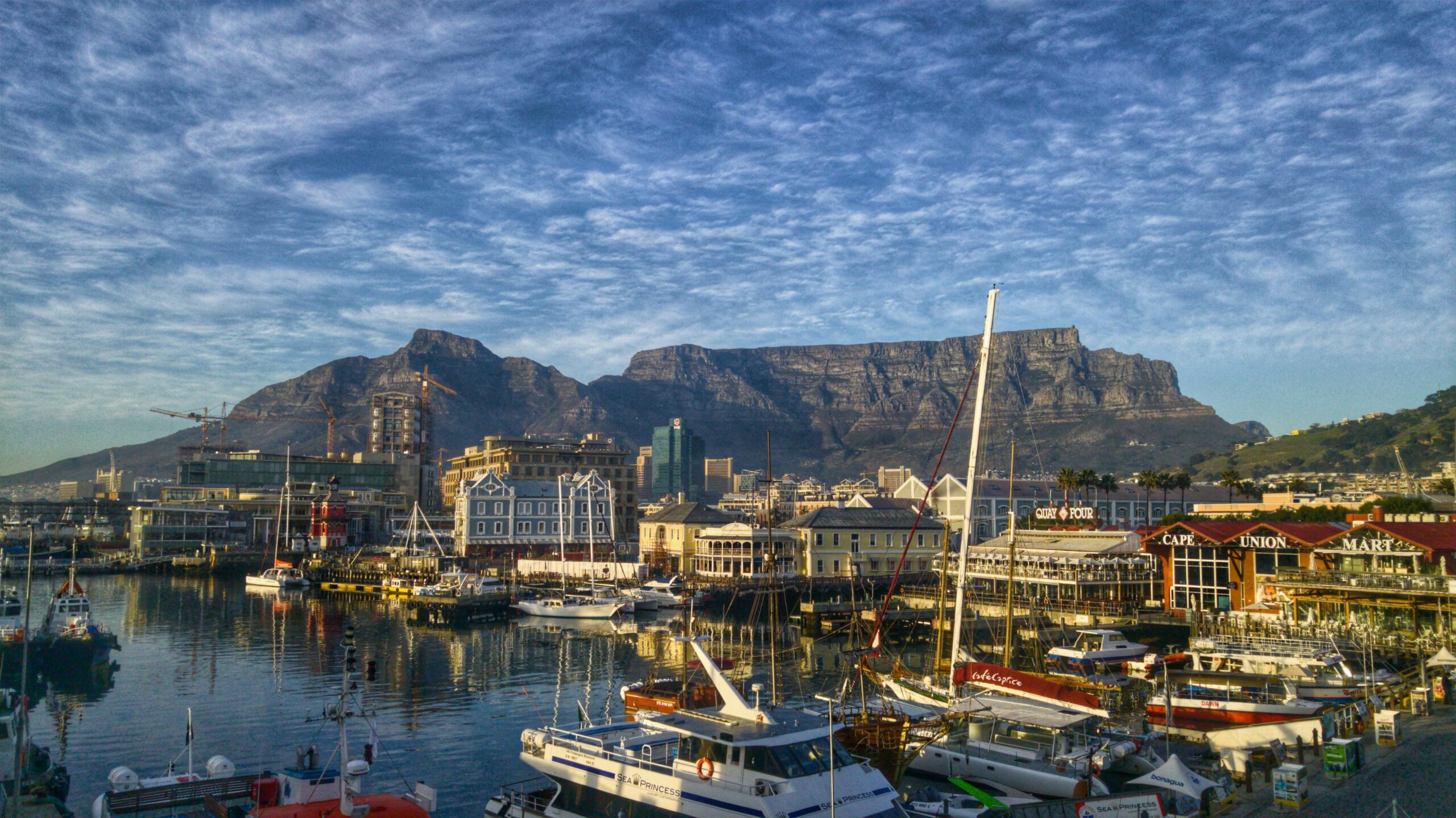 Boats docked at a busy waterfront marina with colorful buildings and shops, set against the backdrop of Table Mountain under a blue, cloud-streaked sky in Cape Town, South Africa.