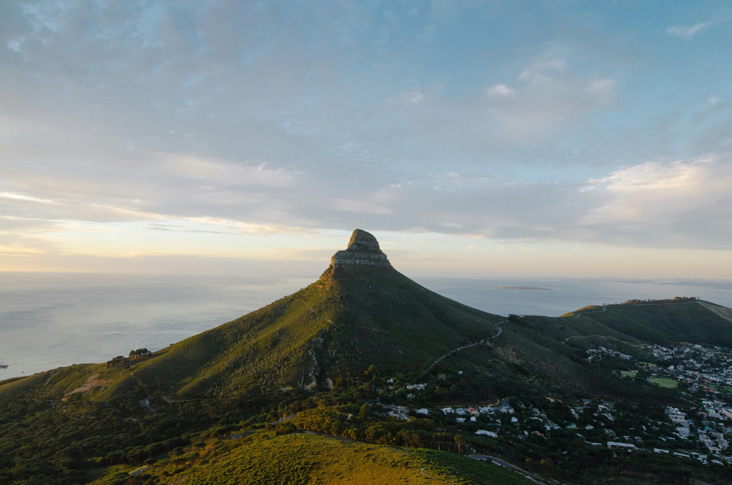 A distinctive, conical mountain rises above green hills, with the ocean and sky in the background. Homes are nestled at the mountain's base, and scattered clouds catch the soft, golden light of sunset.