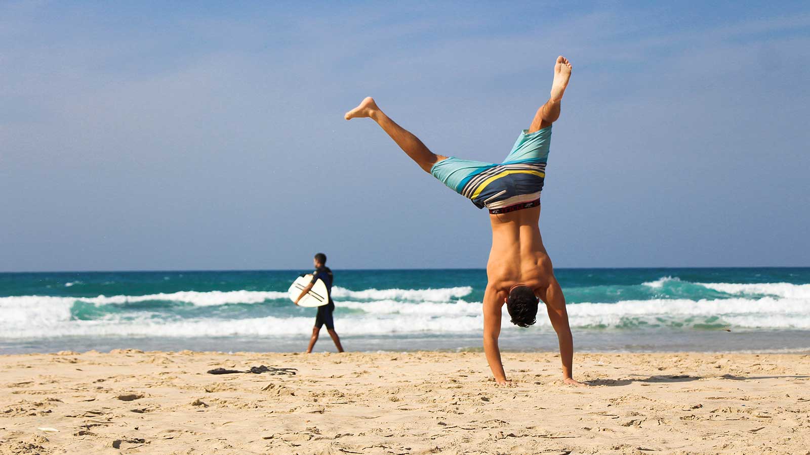 A man does a handstand on a sandy Cape Town beach near the ocean, while another person holding a surfboard walks toward the water in the background—one of many fun things to do here on a budget under the clear blue sky.