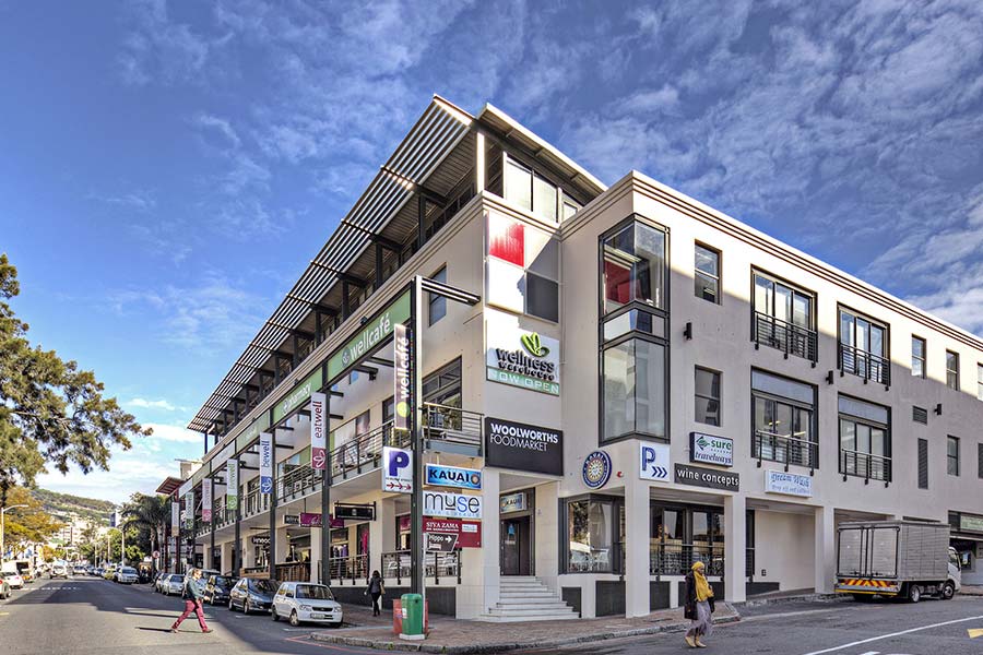 A vibrant, modern three-story commercial building with various business signs and balconies stands on a sunny city street corner, parked cars lining Kloof Street beneath the bright blue sky.