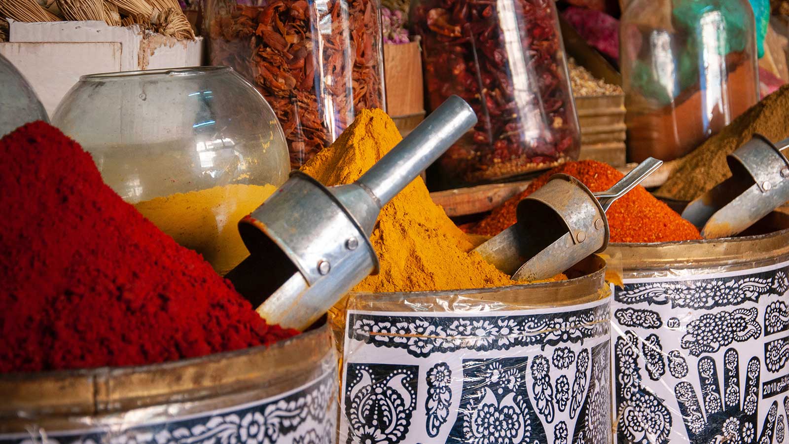 Colorful piles of ground spices—red, yellow, orange, and brown—are displayed in open metal bins with scoops, set against jars of dried herbs in a vibrant market near your holiday accommodation for a memorable stay.