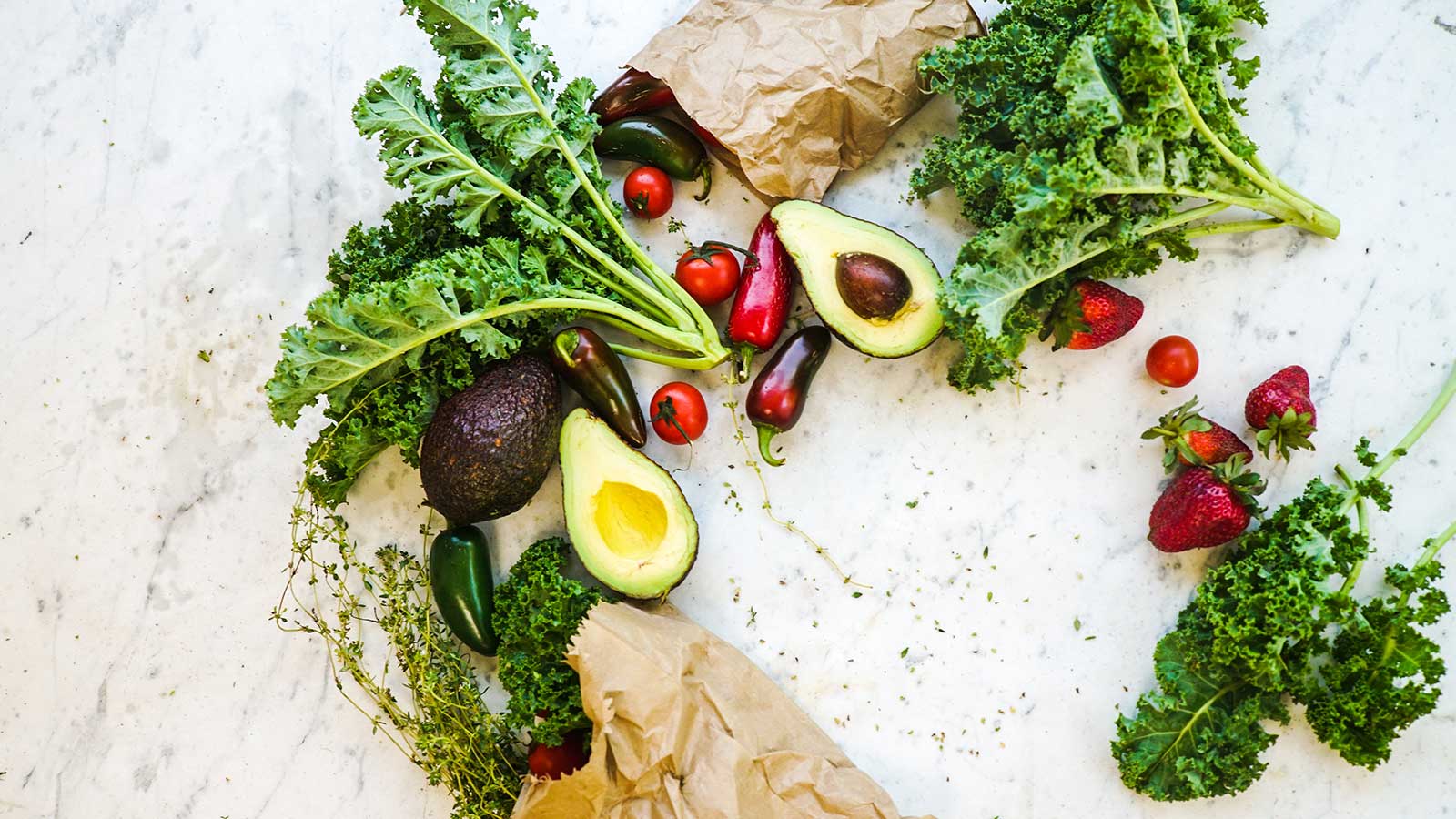 Fresh vegetables and fruits from Gardens Centre—like kale, avocados, cherry tomatoes, and strawberries—are scattered on a white marble surface with brown paper bags, perfect for preparing for your stay at our Holiday Apartments.