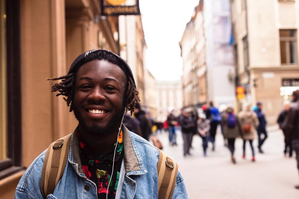 A young man with a backpack and headphones smiles at the camera on a lively city street, capturing the vibrant energy near Gardens Centre Holiday Apartments, an ideal accommodation for students amidst historic buildings and bustling crowds.