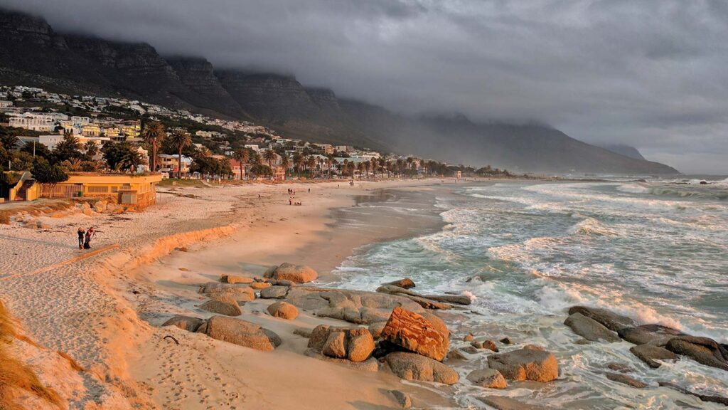 A sandy beach with large rocks, gentle waves, and a cloudy sky; mountains and buildings line the coast near Gardens Apartments, with a few people walking along the shoreline at sunset—one of the best attractions easily reached by My Citi Bus.