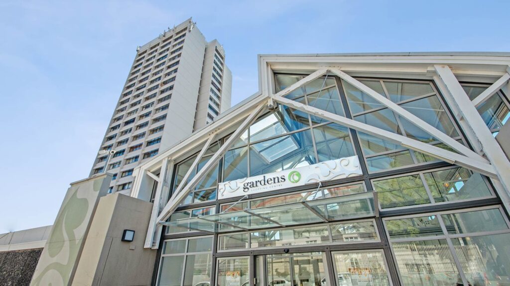 A modern glass and metal entrance labeled "gardens" marks the Gardens Shopping Centre, with a tall white high-rise apartment tower rising in the background against a clear blue sky.