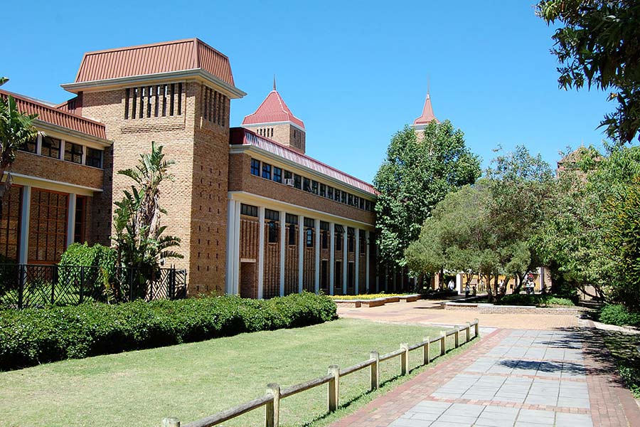 A brick academic building with large windows and red roofs stands beside a paved walkway at the University of Western Cape, surrounded by green grass, bushes, and trees under a clear blue sky.